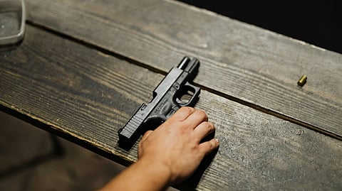A hand reaches for a black handgun on a wooden table, with a single bullet casing nearby. 