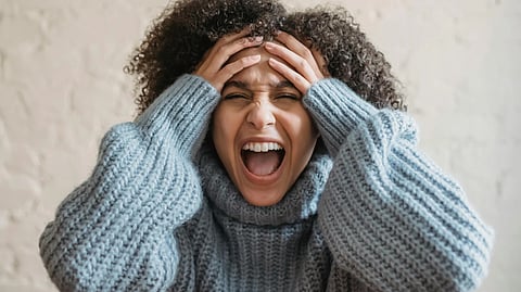 Woman in a gray sweater clutches her head, eyes closed, mouth open, expressing frustration or stress.