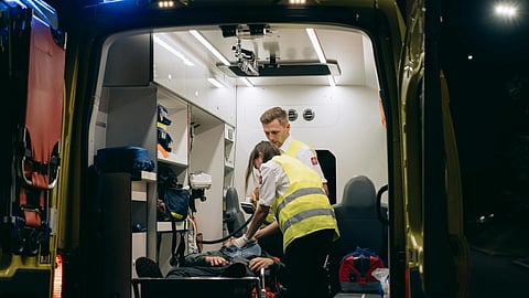 Paramedics treat a patient in an ambulance at night, with blue lights visible. 