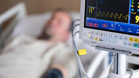 A hospital room showing a blurred patient on a bed with a blood pressure cuff, and a clear monitor displaying vitals.