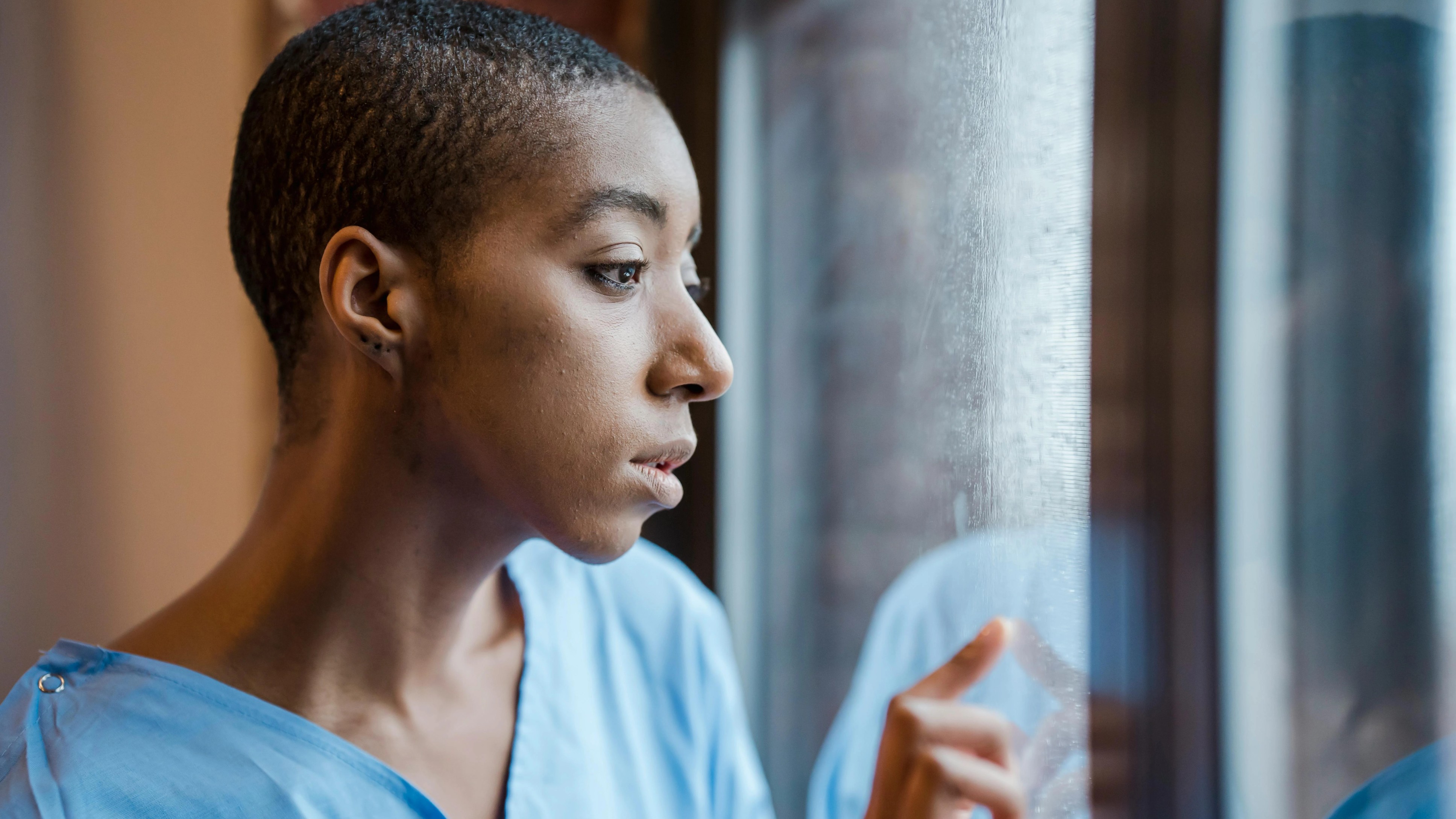 Person in a blue hospital gown gazes pensively out a window, touching the glass lightly.