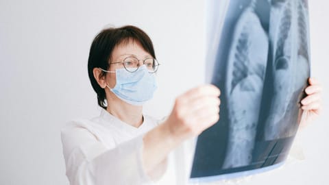 A person in a white coat and mask examines an X-ray image of a chest, appearing focused and analytical. 
