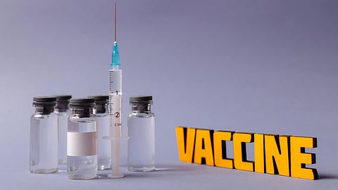 Five vaccine vials and a syringe on a gray surface with “VACCINE” in bold yellow text.