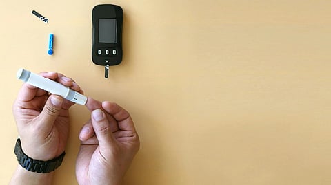 Two hands on a beige background use a white lancing device to test blood sugar. 