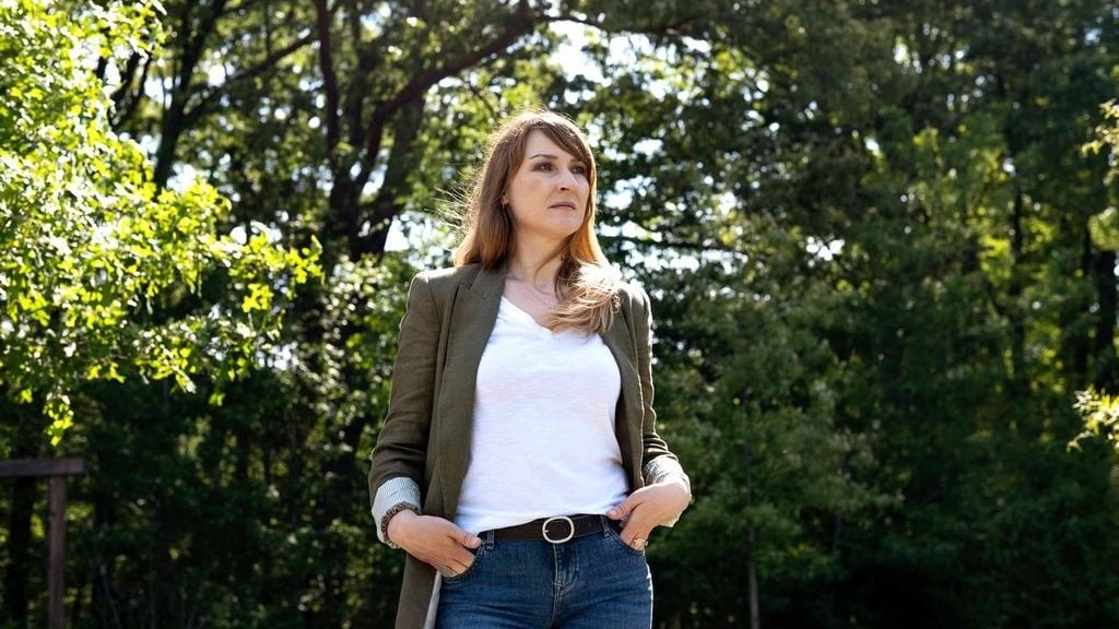 A woman stands confidently outdoors, wearing a white shirt and olive jacket, hands in pockets. 