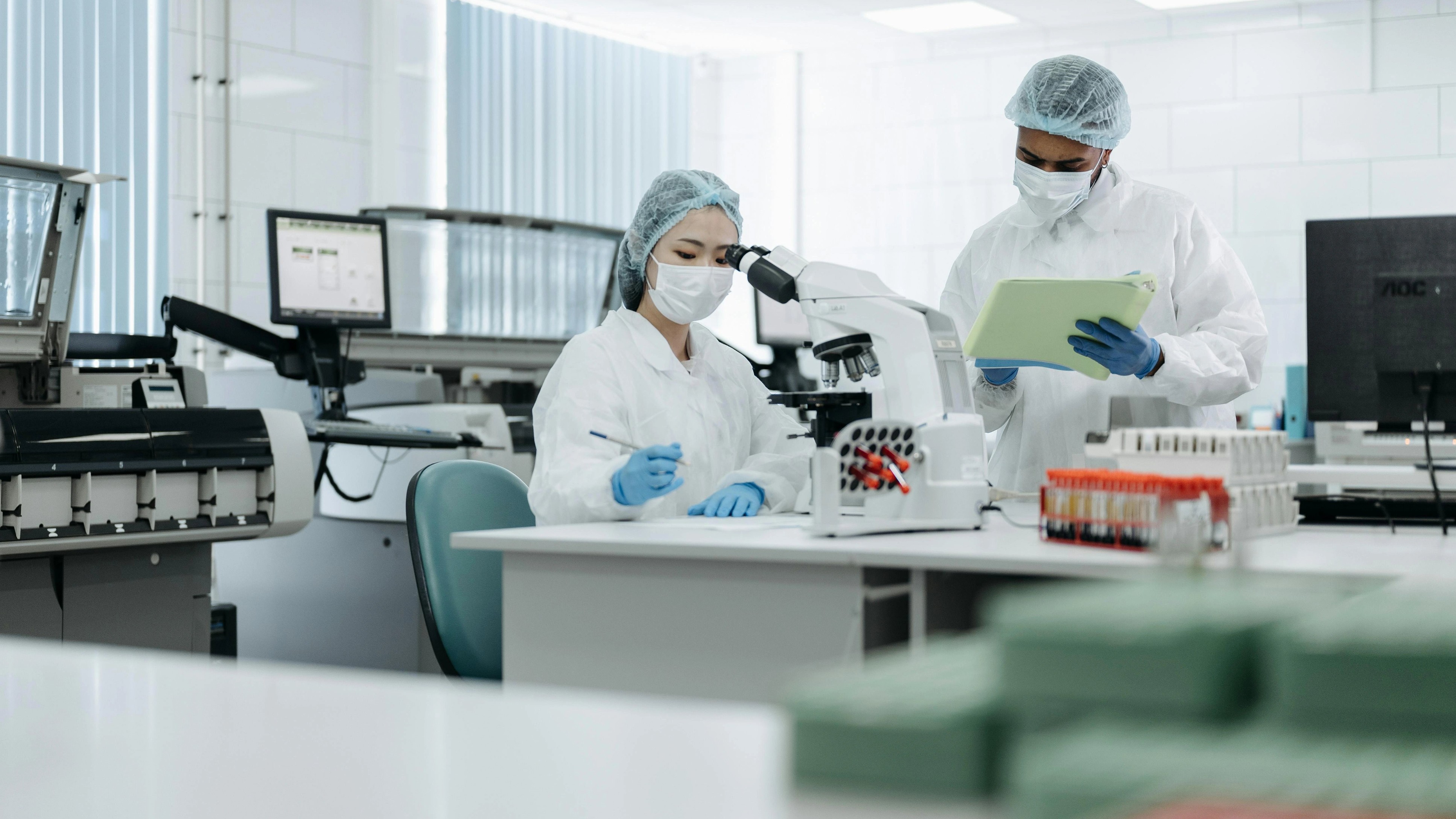 Two scientists in protective gear work in a lab. One uses a microscope, while the other holds a clipboard.