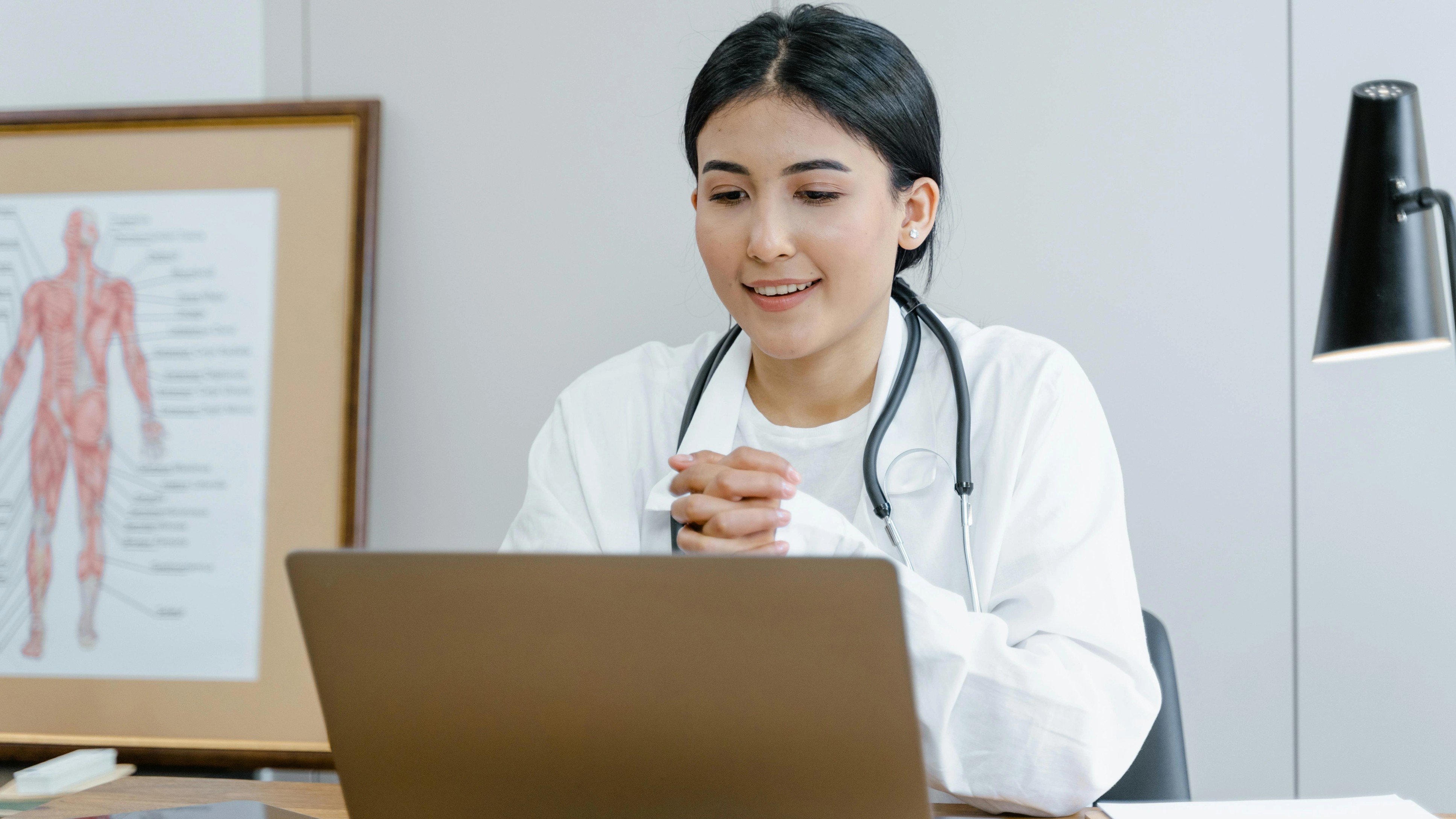 A doctor in a white coat with a stethoscope smiles while looking at a laptop.