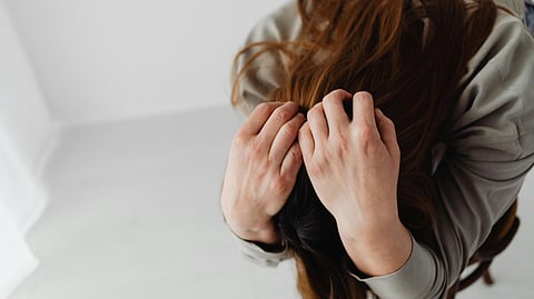 A person with long brown hair is bent over, clutching their head with both hands. 