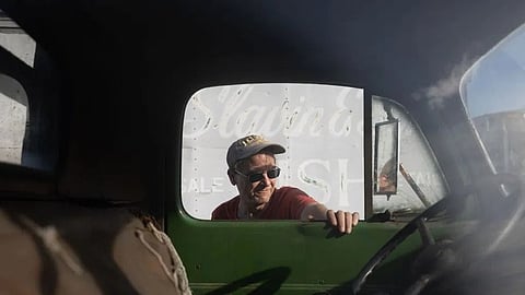 A man in a cap and sunglasses leans on a green vintage truck's door, partially shadowed. 
