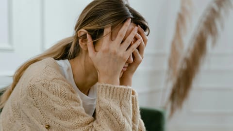 A woman in a beige sweater sits in a room, holding her head in her hands.