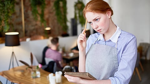 A café server in an apron holds a notepad and pen while thinking about an order.