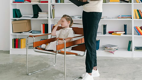 A child sits in a chair with a hand on their forehead, appearing thoughtful or puzzled.