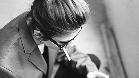 A black and white photo of a person with glasses, bun hairstyle, and a cigarette, wearing a blazer.