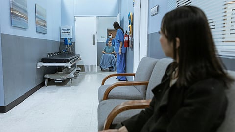 A hospital hallway with light blue walls. A person sits on a couch in the foreground.