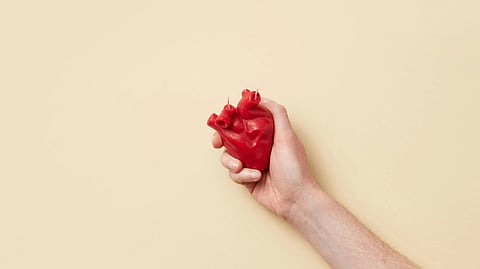 A hand holds a red candle shaped like an anatomical heart against a beige background.