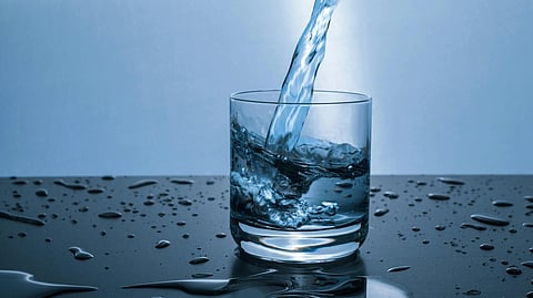 A stream of water pours into a clear glass, causing splashes on a dark, wet surface. 