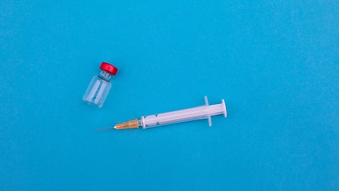 A syringe with an orange needle lies next to a small transparent vial with a red cap on a blue background.
