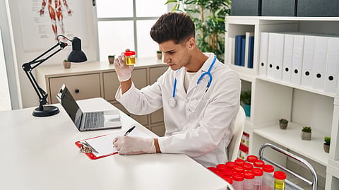 A male doctor examines a urine sample in a bright office. He's writing notes on a clipboard.