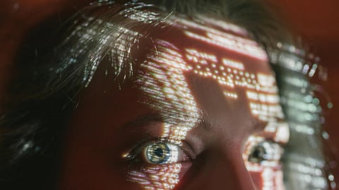A face with projected light patterns and symbols, highlighting expressive wide-open eyes. 