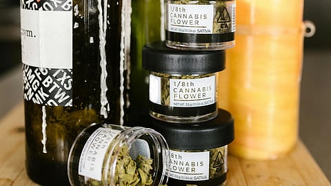 Wine bottles with wax drips, stacked cannabis jars, and a lit candle are arranged on a wooden surface.