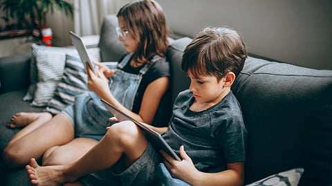 Two children, one in glasses, sit on a gray sofa using tablets. 