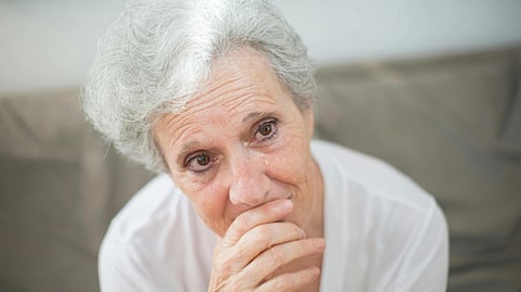 An elderly woman with short white hair, wearing a white shirt, sits on a sofa and looks thoughtfully with her hand on her chin.