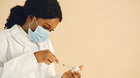 A healthcare professional in a mask and lab coat draws liquid from a vial with a syringe. 