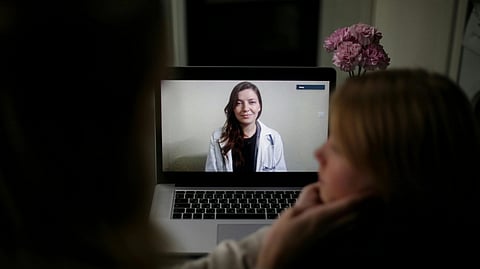 A woman sits at a laptop in a dimly lit room, engaged in a video call with a doctor. 