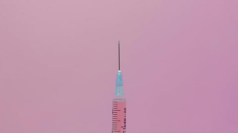Close-up of a syringe with a blue cap and needle against a pink background.