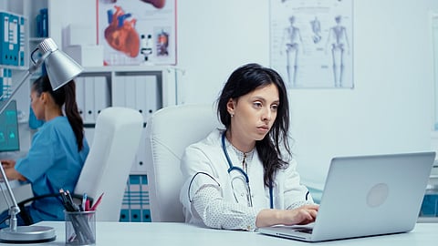 Young woman doctor in private medical cabinet typing on laptop while the nurse in working in the background