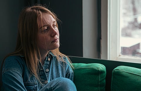 A sad young woman is sitting on the sofa by the window