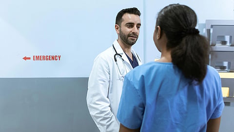 A doctor in a white coat talks to a nurse in blue scrubs in a hospital hallway.