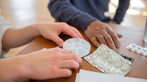 Two individuals sit at a table with medication packs, a pill organizer, and a notepad. 