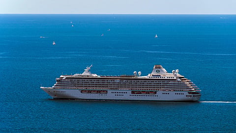 A large, white cruise ship sails through calm, deep blue ocean waters under a clear sky.