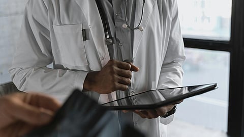 A doctor in a white coat and stethoscope holds a tablet indoors, focusing on the screen. 