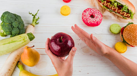Hands hold a red apple, rejecting junk food. Surrounding are fruits, vegetables, a hot dog, and a donut on a wooden background.