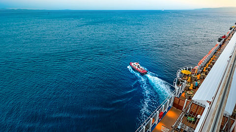 Cruise ship deck overlooking vast blue ocean, with a small red boat creating a white trail. 