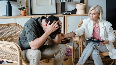 A man sits with his head in his hands, visibly distressed. A woman sitting beside him gently touches his arm.