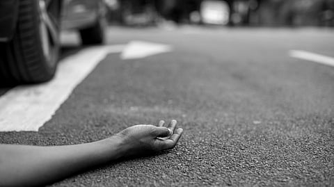 A hand lies motionless on a city street near a car's rear wheel. 