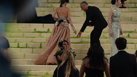A woman in a voluminous pink gown ascends grand steps, assisted by a man in a suit. 