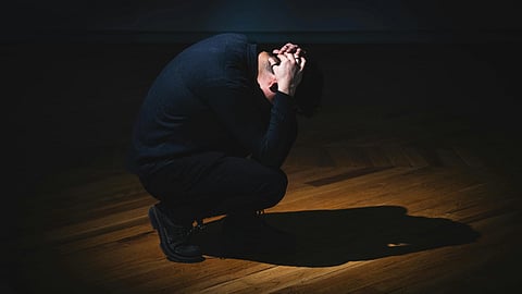 A person crouched on a wooden floor, hands clasped over their head, under a spotlight.