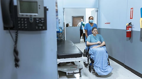 A nurse in scrubs and mask pushes a patient in a wheelchair through a sterile hospital hallway.