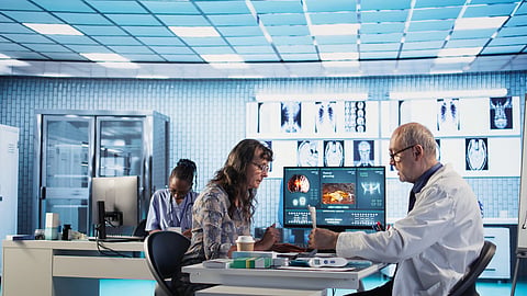Two doctors review medical scans on a computer in a lab while another works at a desk.