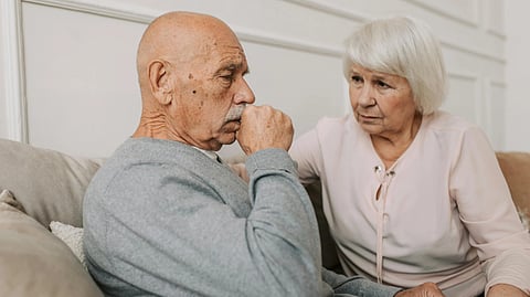 An elderly man coughing on a sofa while a worried woman sits beside him.