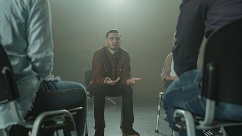 A man in a brown jacket speaks earnestly in a support group setting, seated in a circle.