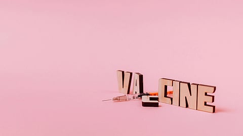 Wooden letters spelling “vaccine” separated by a syringe on a pink background.