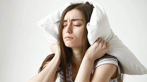 A woman with long hair squeezes a pillow over her ears, eyes closed and frowning.