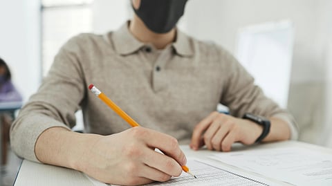 A person in brown shirt and mask, taking an exam, holding a pencil.