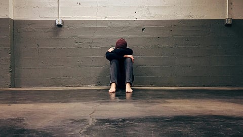 Person in a beanie sits with their head on knees in a corner of a bare concrete room, barefoot.