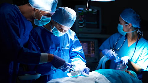 Surgeons in blue scrubs and masks performing an operation under bright lights.